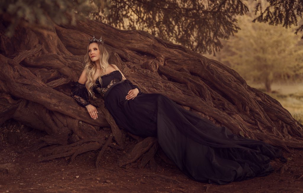 dreamy outdoors maternity photo pregnant lady laying on and old tree roots dressed in black dramatic gown Surrey maternity photoshoot