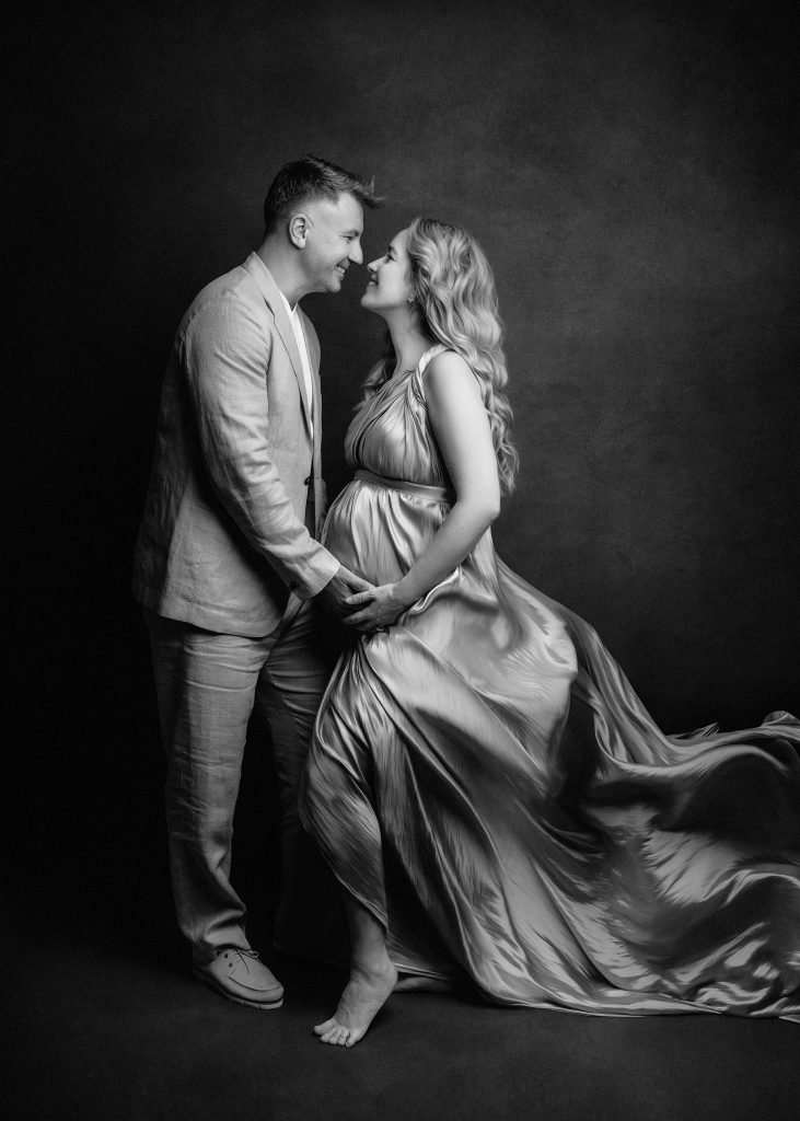 black and white maternity photo with a partner studio classic mood standing facing each other wind blows the pregnancy gown smiling enjoying natural pose Surrey photoshoot