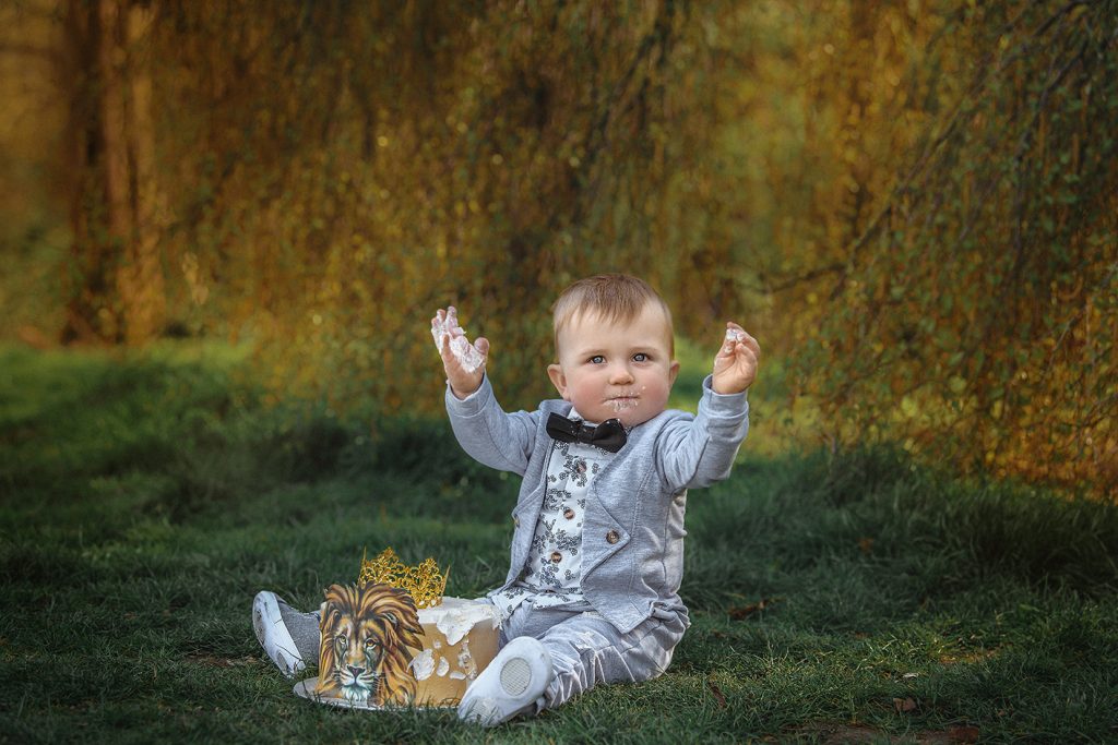 Baby first birthday cake smash outdoors photoshoot baby sitting on the grass in the park clapping hands smashing the cake Cheam Sutton Surrey South London photoshoot