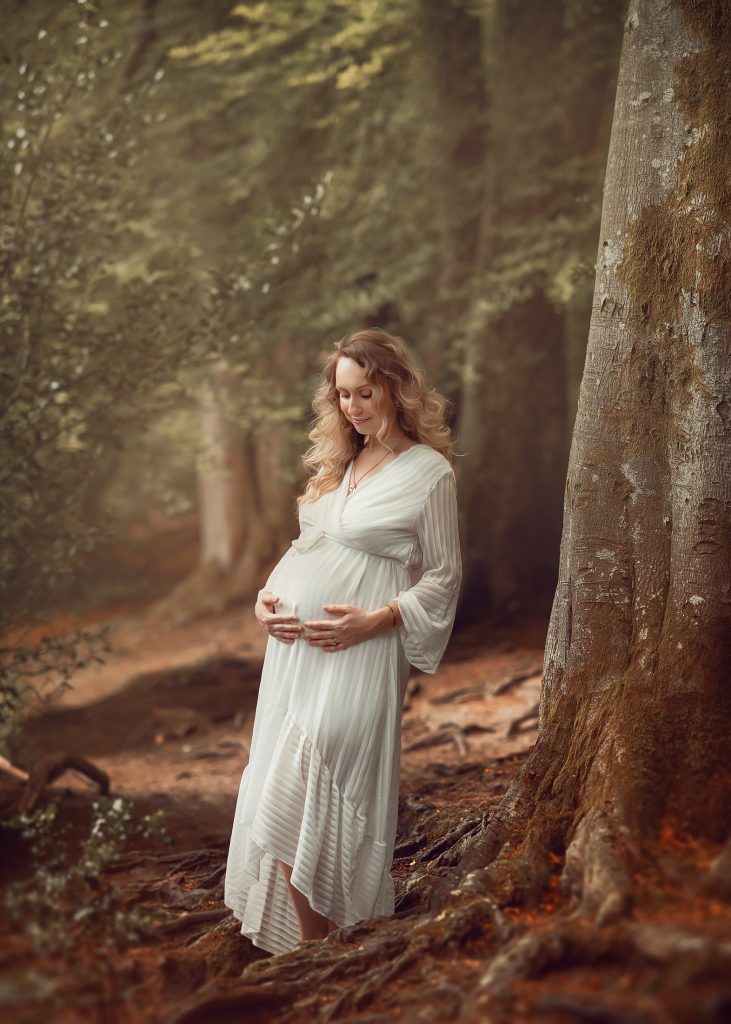 natural maternity photo wearing casual long white dress woods surroundings happy mood holding belly looking down Headley Box Hill pregnancy photoshoot Surrey