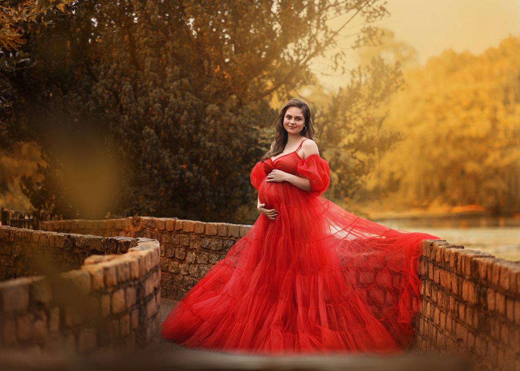 maternity dreamy photo taken in Beddington Park Wallington pregnant lady wearing long tulle red dress posing on a stone bridge classic holding belly pose natural pregnancy photo