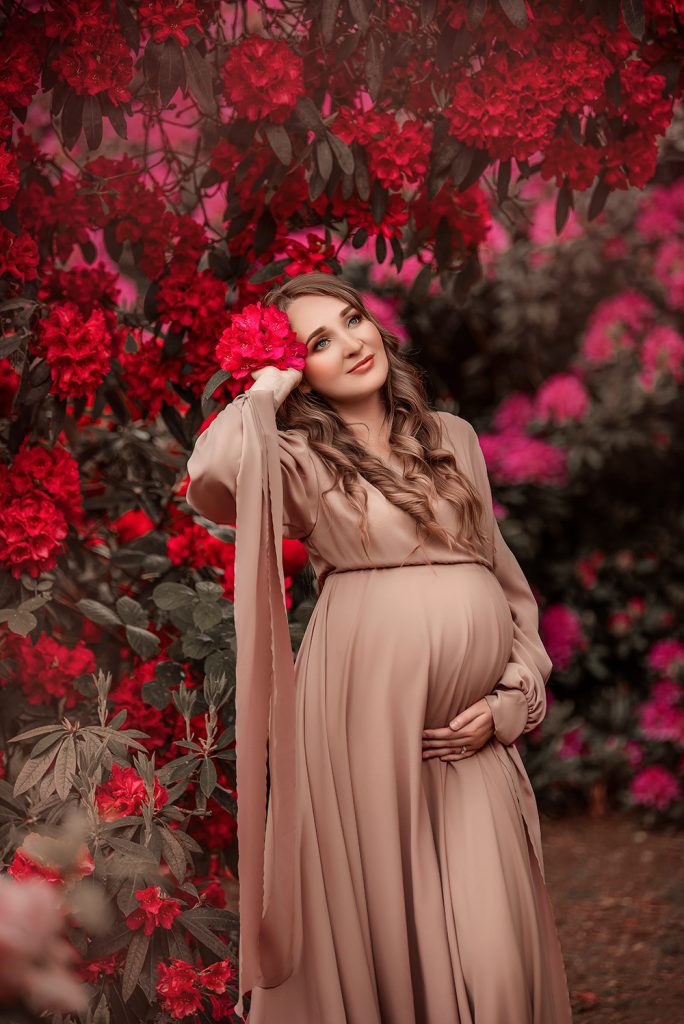 Creative Maternity Photography Outdoors pregnant lady surrounded by blooming bushes posing in natural way holding belly wearing soft rosy gown London South London photoshoot