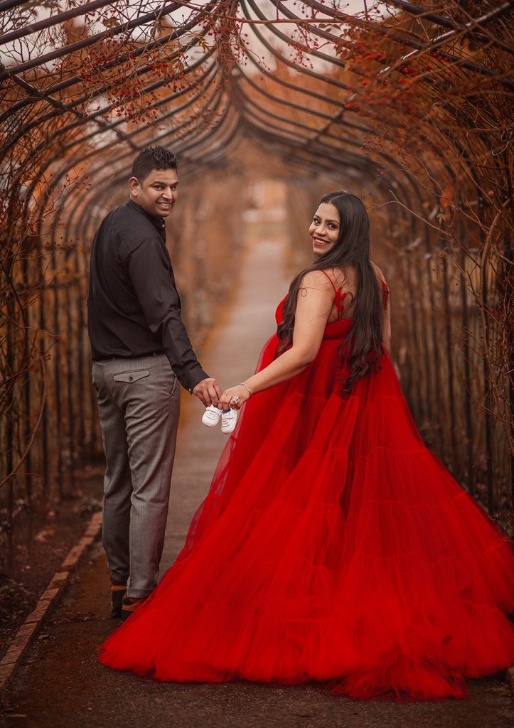 pregnant couple posing in the park holding small baby shoes together facing camera smiling lady wering long red pregnancy dress Cheam Sutton Surrey photoshoot