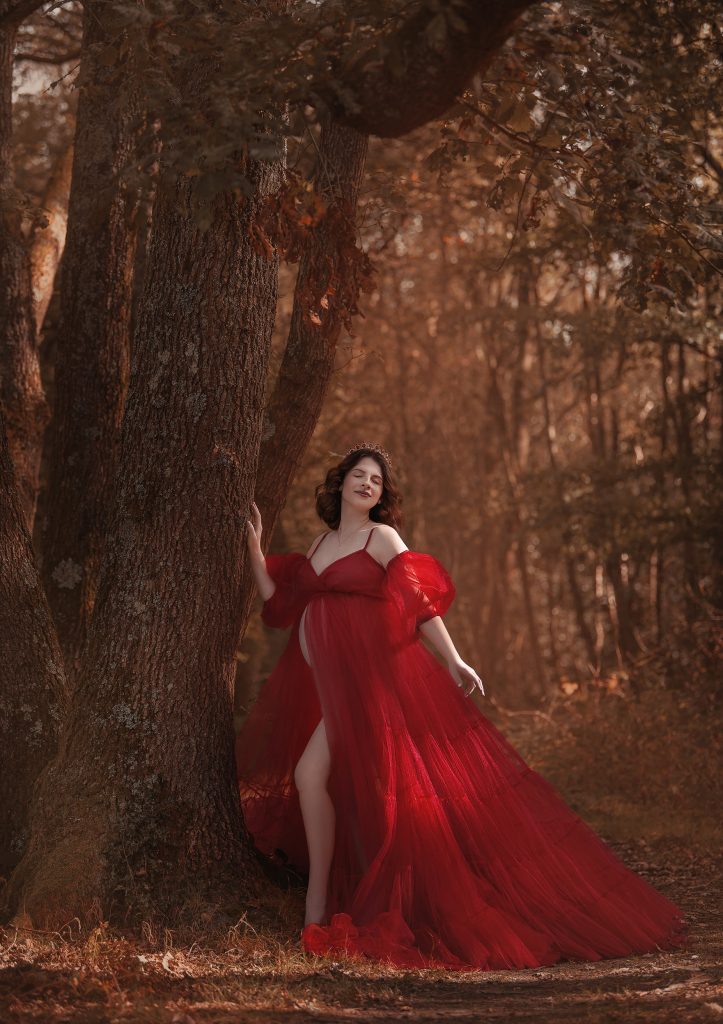 magical outdoors maternity portrait lady wearing red big tulle dress standing in the woods dreamy mood elegant pose warm colours pregnancy photoshoot in Sutton London and Surrey