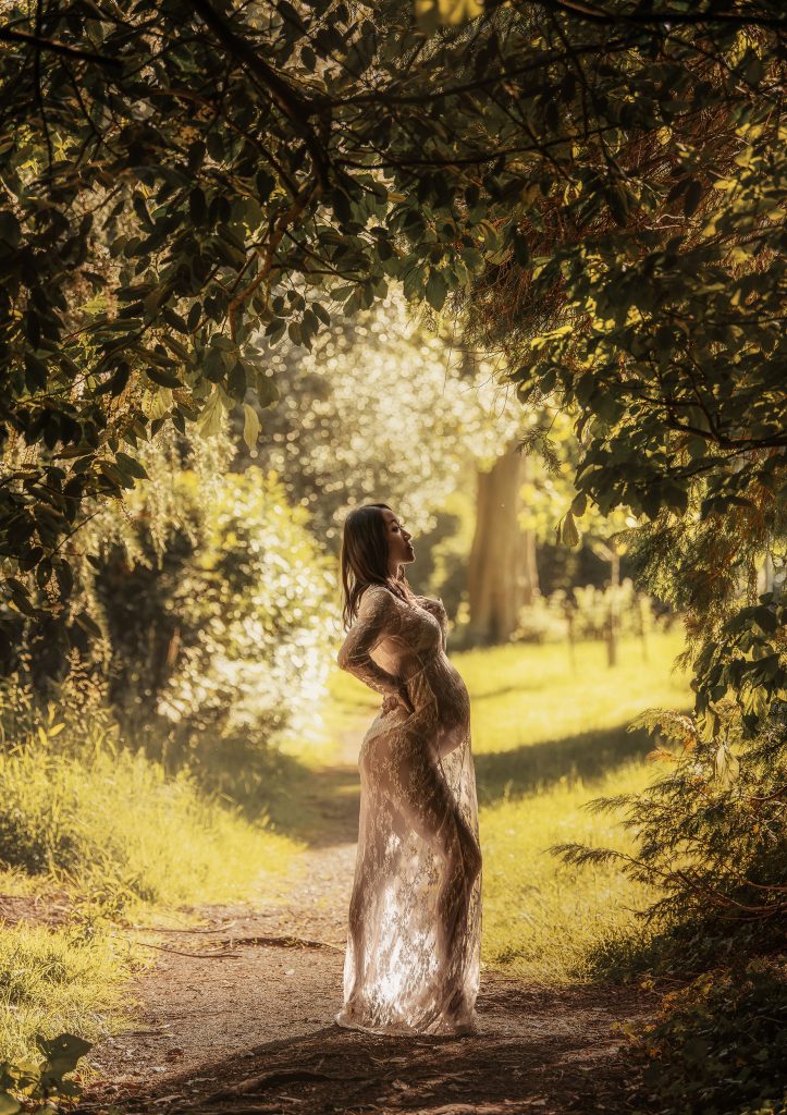 outdoors natural maternity photo pregnant lady standing holds belly wearing lace gown in front of the trees backlit photo in Cheam Sutton South London