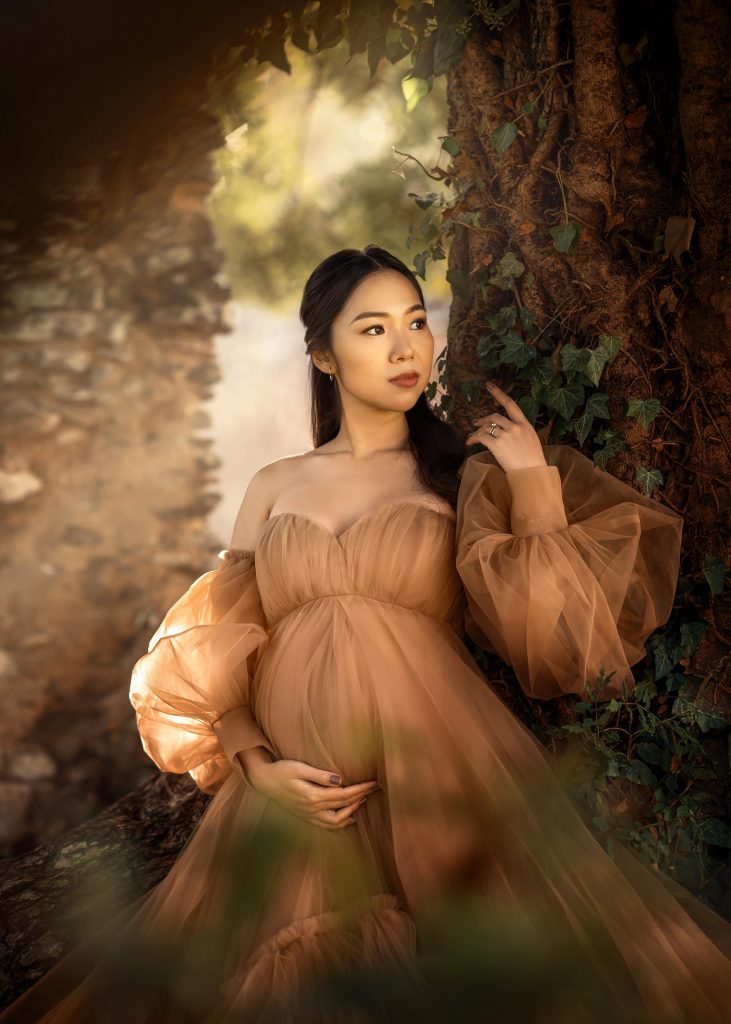 outdoors dreamy maternity portrait pregnant lady standing in between ruins and trees holding belly wearing soft tulle beige dress Surrey photoshoot