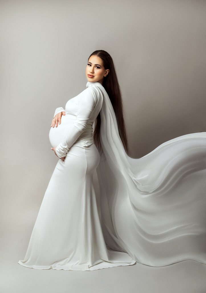 classic elegant studio maternity portrait lady standing in a side pose holding belly wearing white long dress flowing fabric draped from her shoulders down shot taken in Sutton South London and Surrey