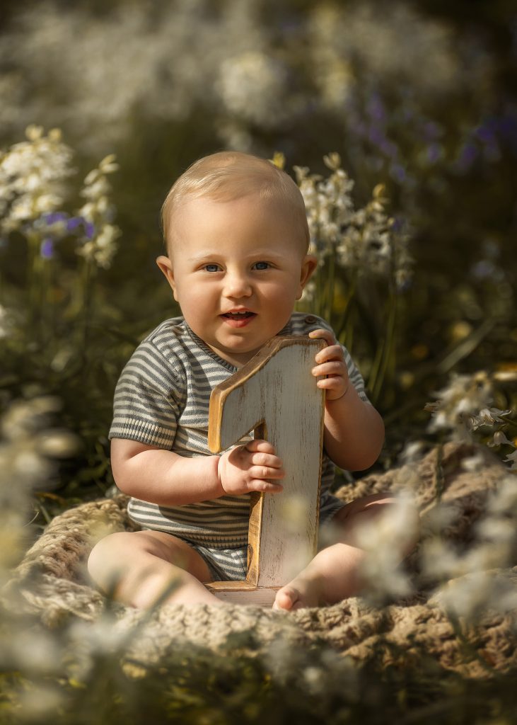 baby boy forst birthday outdoors portrait baby sitting in between flower meadow holding wooden number one smiling Cheam Sutton Carshalton Wallington Croydon photoshoot