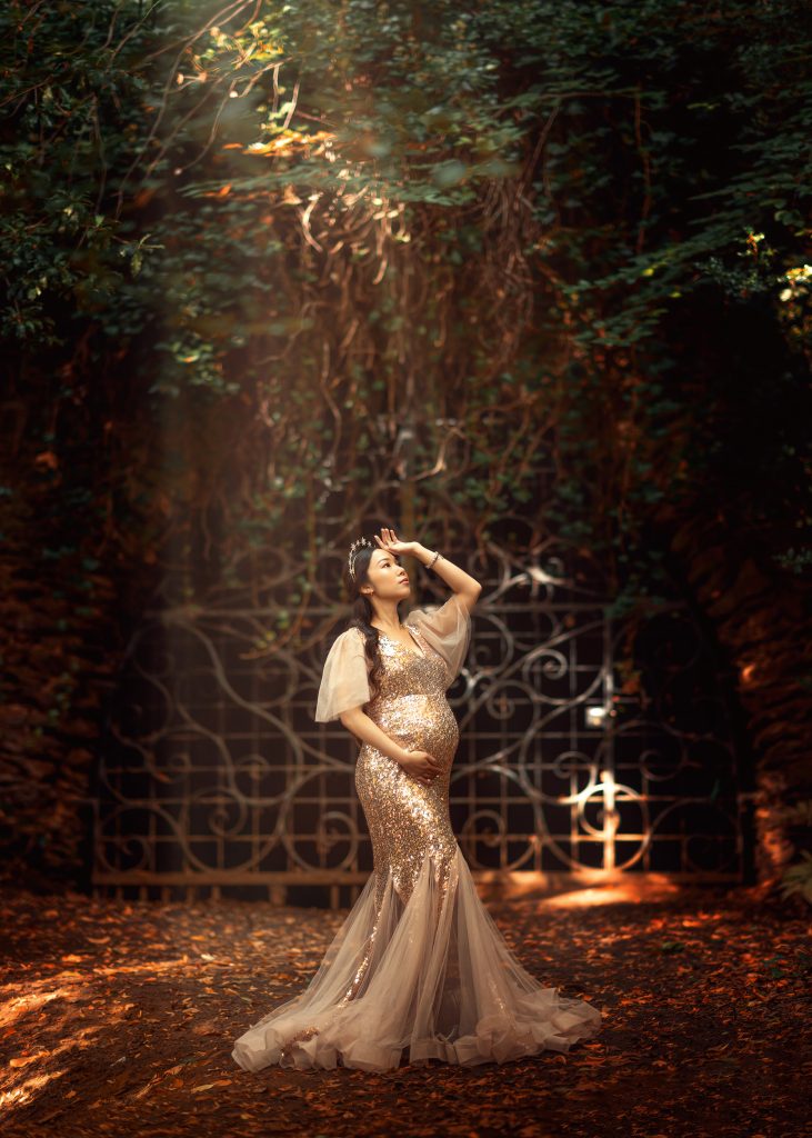 dreamy goddess style maternity photo in Surrey pregnant lady facing the light standing in dramatic wood surroundings wearing long golden dress