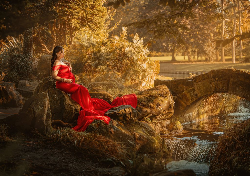 golden hour maternity photo taken in Beddington Park, Wallington pregnant lady sitting o the stones wearing long red dress enjoying the moment