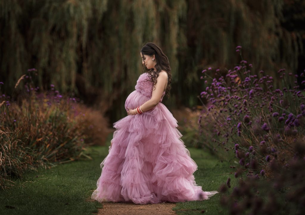 maternity photo pregnant lady standing o the side holds belly wearing pink ruffled long dress summer time at Beddington Park, Wallington photography
