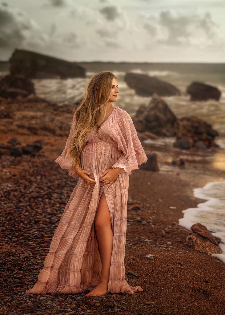 pregnant lady standing at rocky beach wearing long rose bohemian gown looking at the sea smiling and holding belly dreamy natural pose Surrey maternity photoshoot