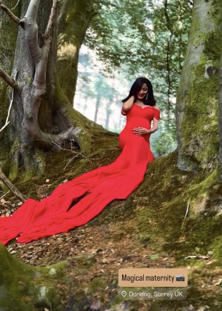 pregnant lady sitting in between old tress in Surrey woods holding belly guided posing stress free wearing long red dress