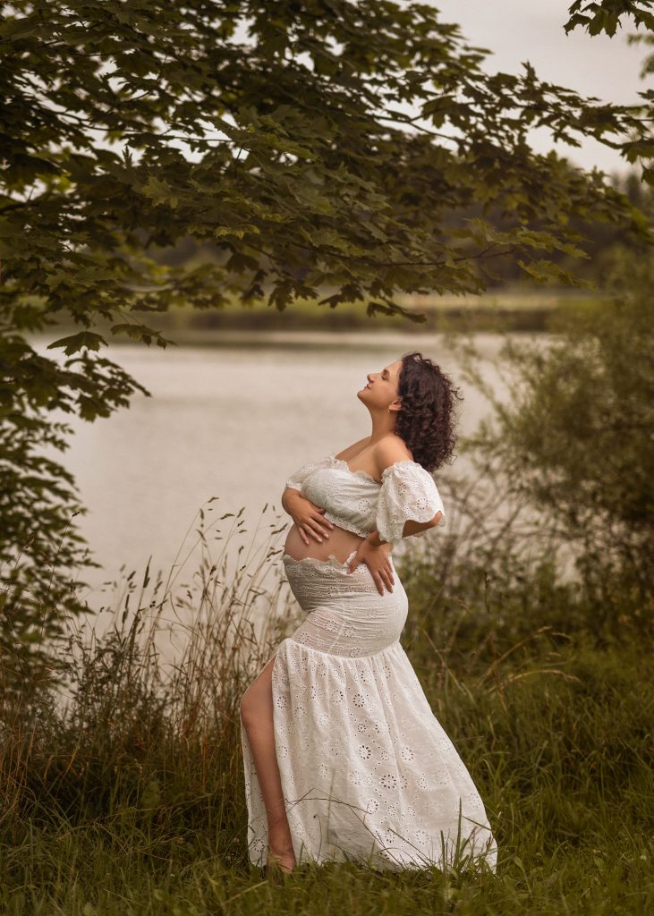 expectant pregnant lady standing on the side holding belly in white outfit posing surrounded by trees in the background of the river